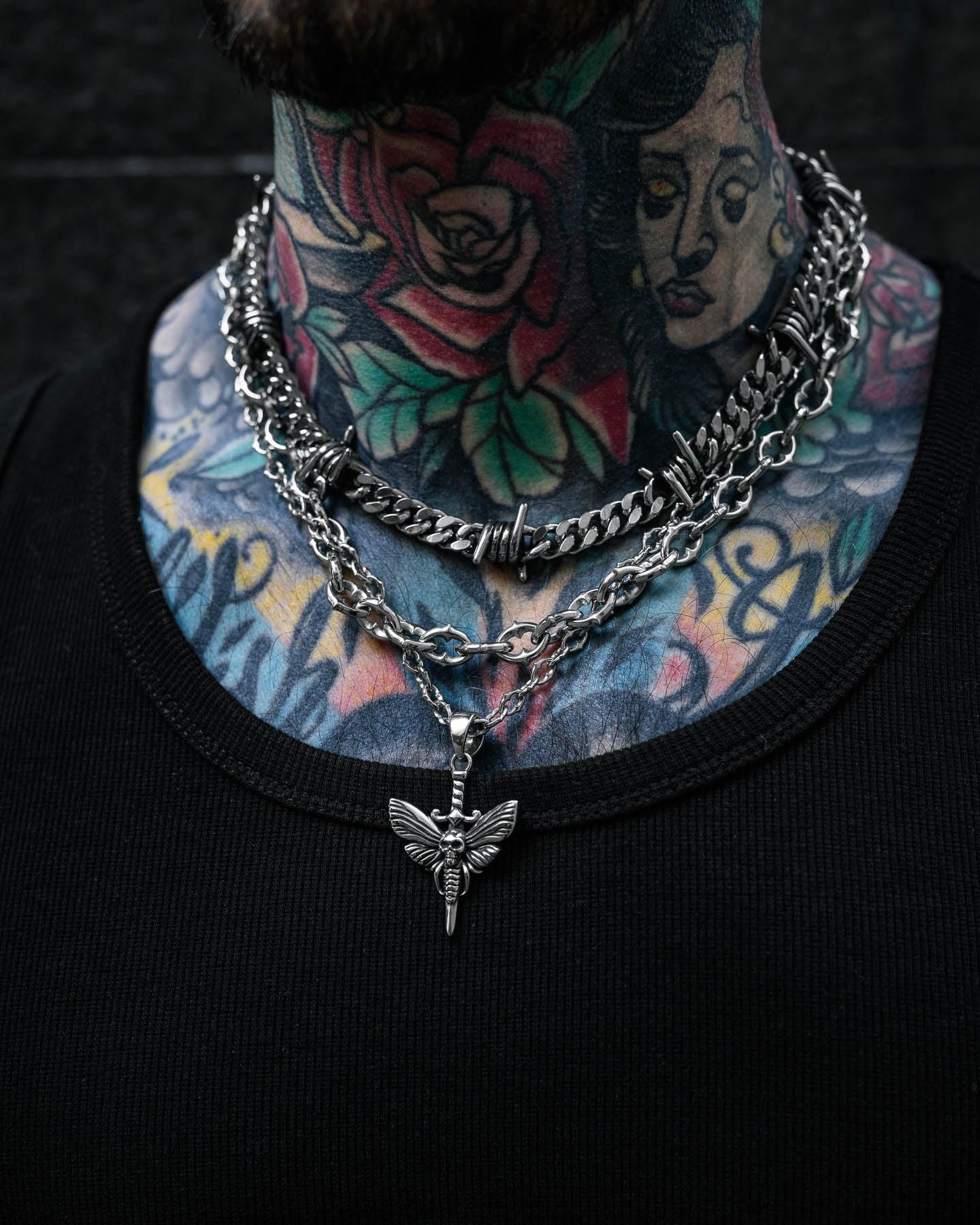 A person with colorful rose and woman's face neck tattoos wears layered silver chains, including the Spite Stack (Custom Pendant Choice) necklace with a spiked chain and moth pendant, paired with a black ribbed shirt.