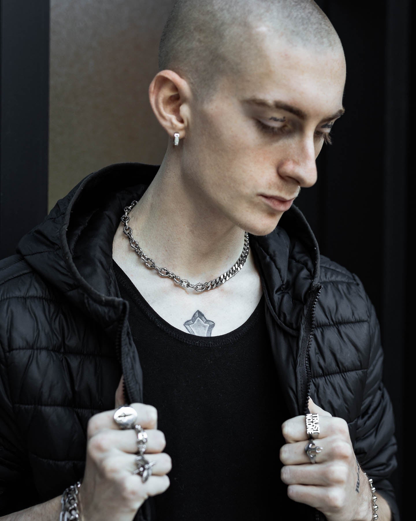 A young person with a shaved head wears a black jacket and shirt, layered gothic jewelry, and the "Split" Cathedral Cuban Chain (Adjustable). They hold their jacket collar, revealing a star chest tattoo and looking slightly downward.