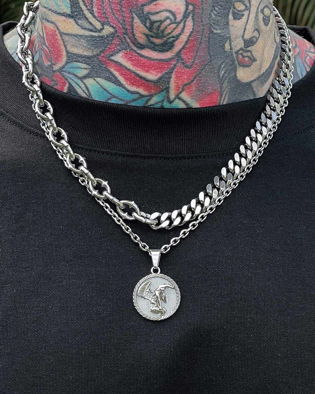 A person with colorful neck tattoos wears gothic jewelry—a spiked chain and the "Split" Cathedral Cuban Chain (Adjustable) with a coin pendant showing a winged figure—over a black shirt. Another chain has a star-shaped design.