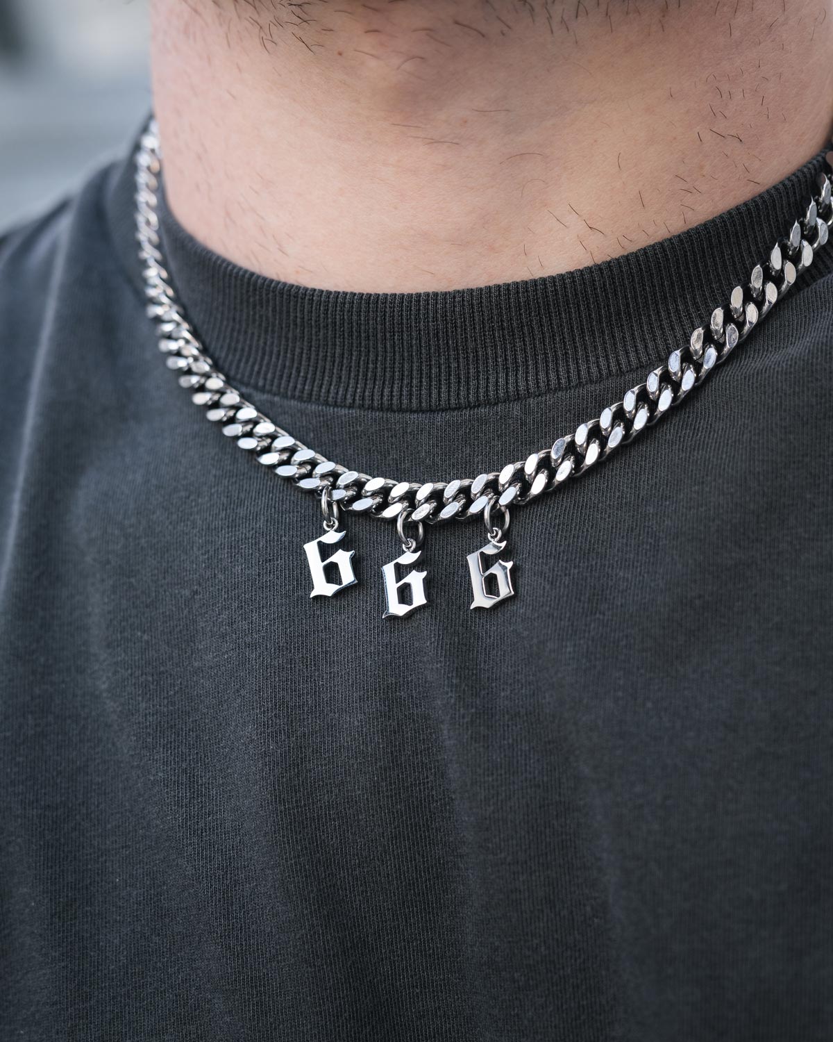 A person wears the Statement Collective Statement Piece Customisable Necklace, a chunky silver Cuban link choker with three metallic pendants shaped as stylized number 6s, over a plain dark gray t-shirt.