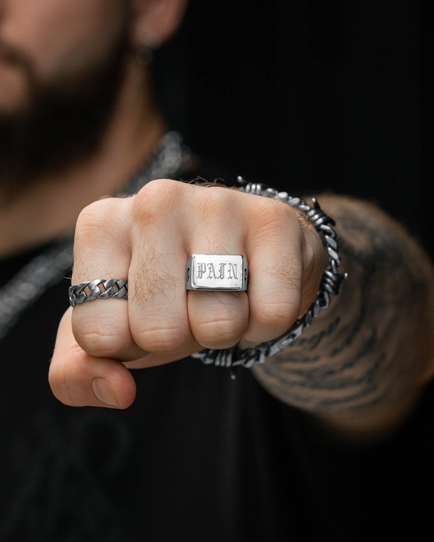 A close-up of a clenched fist wearing the Statement Signet Ring, engraved with "PAIN." The tattooed hand also features a chain bracelet and another ring, with a dark, blurred background highlighting the bold style.
