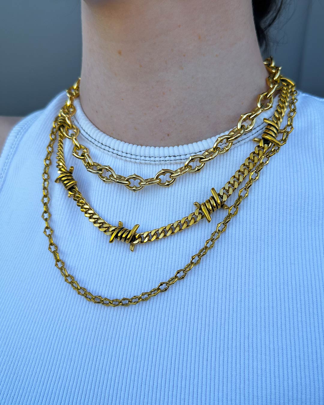 A person wears a white ribbed top with the Stay Safe Stack (Gold), a layered gold necklace set featuring barbed wire and spiked chain styles.