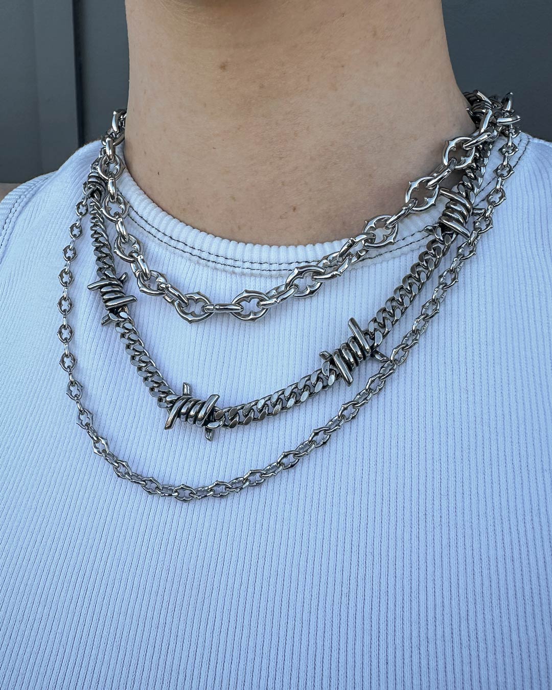 A person in a white ribbed shirt showcases the Stay Safe Stack, featuring bold silver-toned chunky chains and a striking barbed wire necklace layered around their neck.