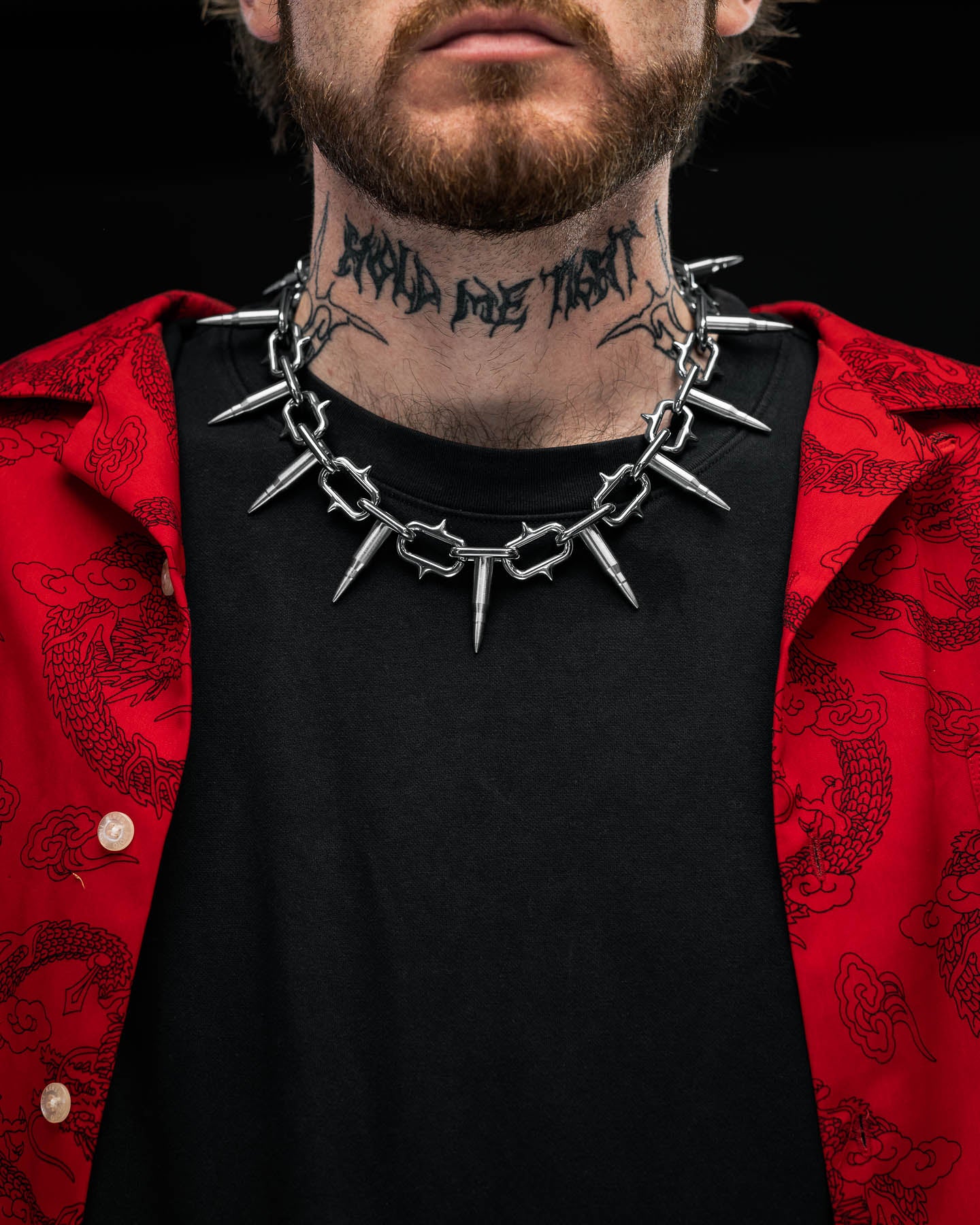 A person wearing a red patterned shirt over a black t-shirt, accessorized with the "Sweating Bullets" Necklace Chain by Statement Collective and a Hold the Trust neck tattoo. The image is cropped to show only their lower face and chest.