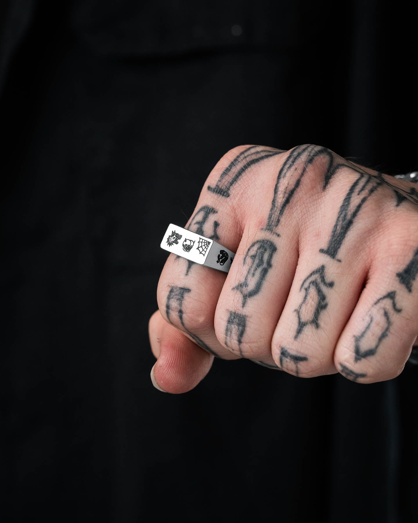 A hand with bold tattoos wears the Tattoo Signet Ring—a stainless steel signet engraved with sun, shield, and fist symbols—on the middle finger. The background is dark and blurred.