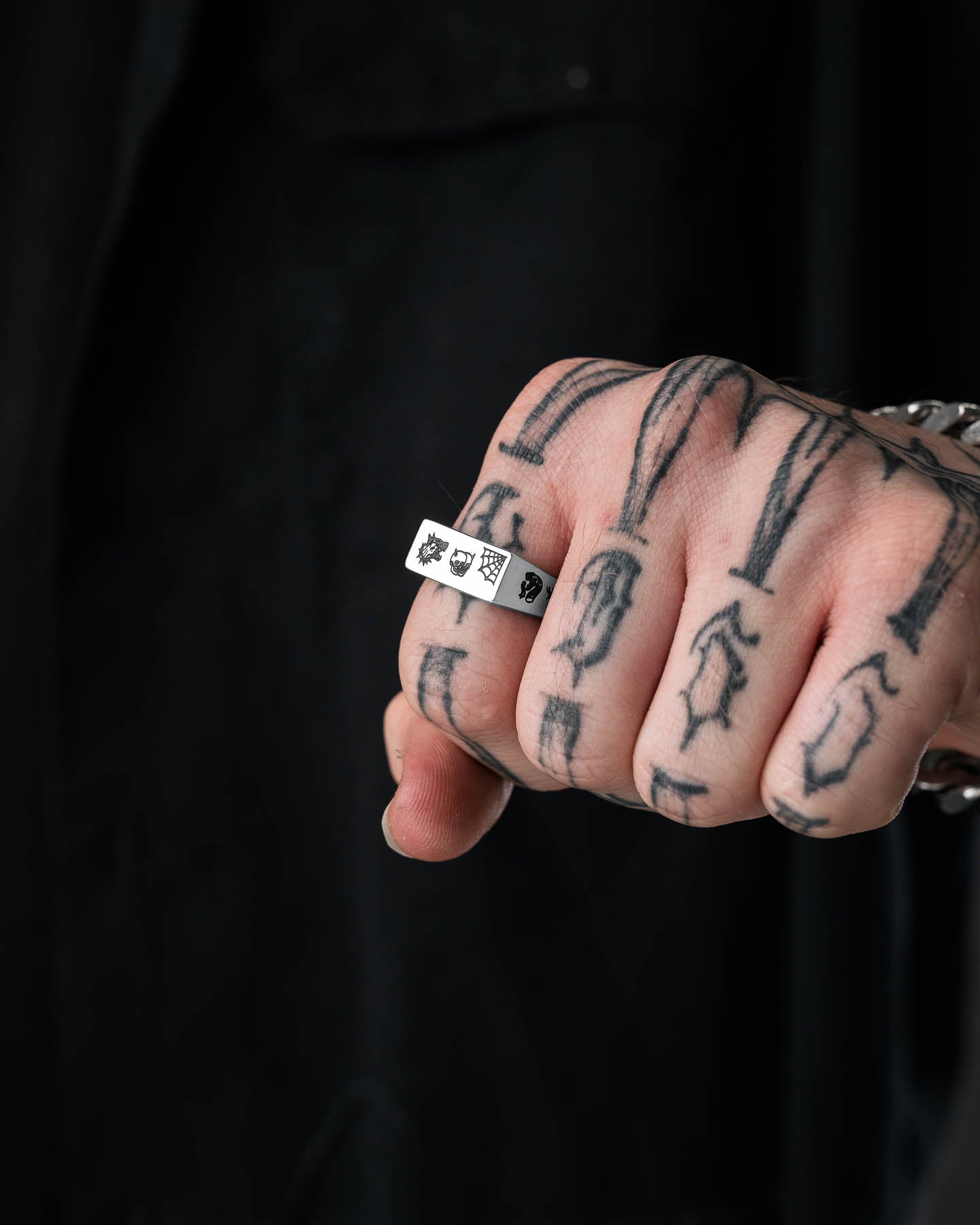 A close-up of a tattooed hand wearing the Tattoo Signet Ring, crafted in stainless steel and featuring playing card symbols, set against a dark background with the person dressed in black.