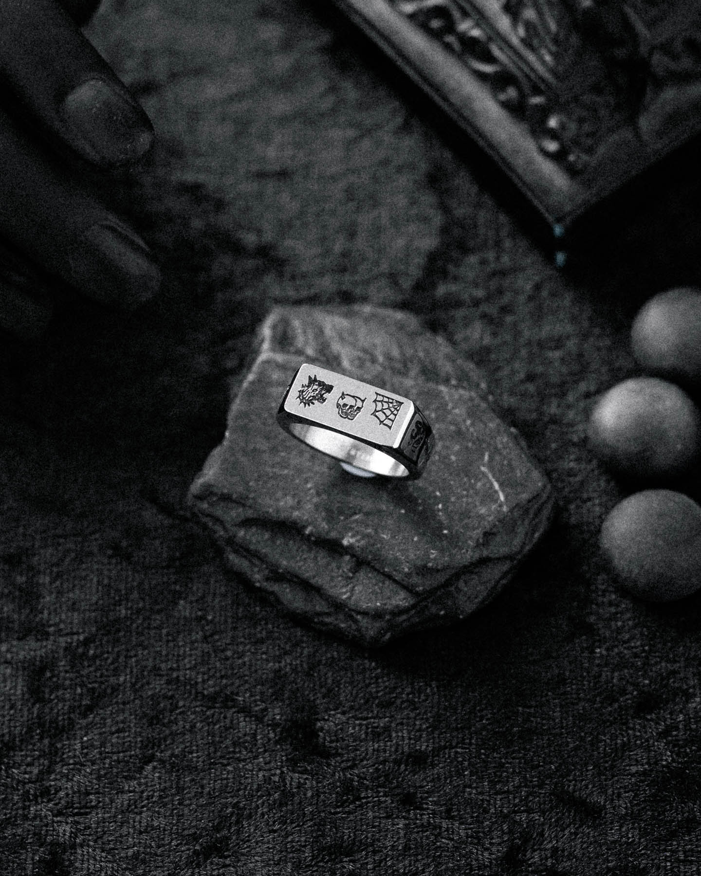 The Tattoo Signet Ring, featuring engraved symbols on silver, rests on a rough stone slab amid round accents and a partially seen hand, all set against a textured dark background.