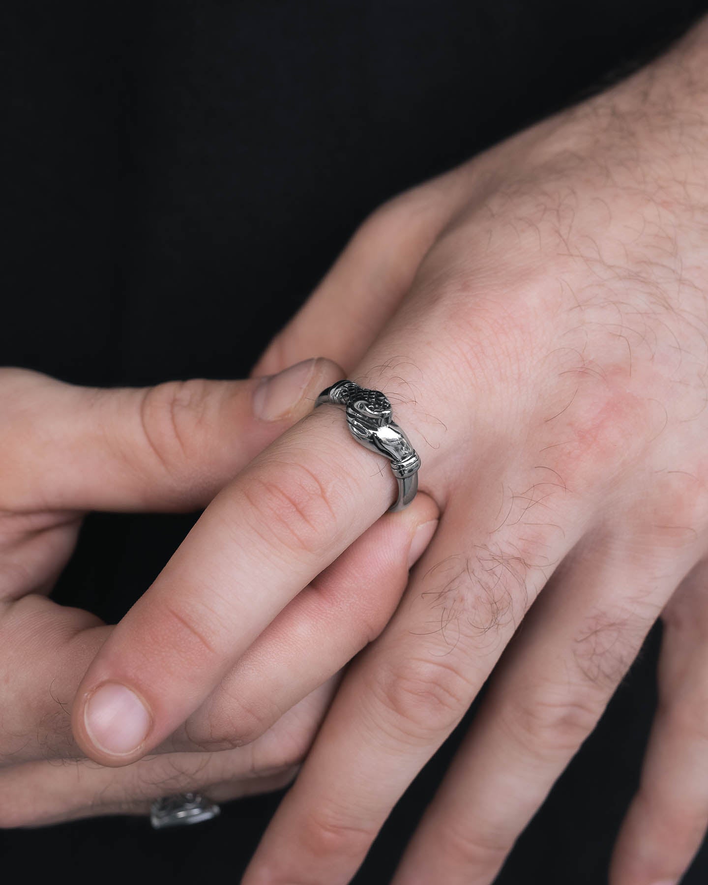 A close-up of a hand wearing "The Hand That Bites" Ring, a unique stainless steel piece with a snakeskin pattern. The other hand gently touches the ring finger against a black background, highlighting the striking design.
