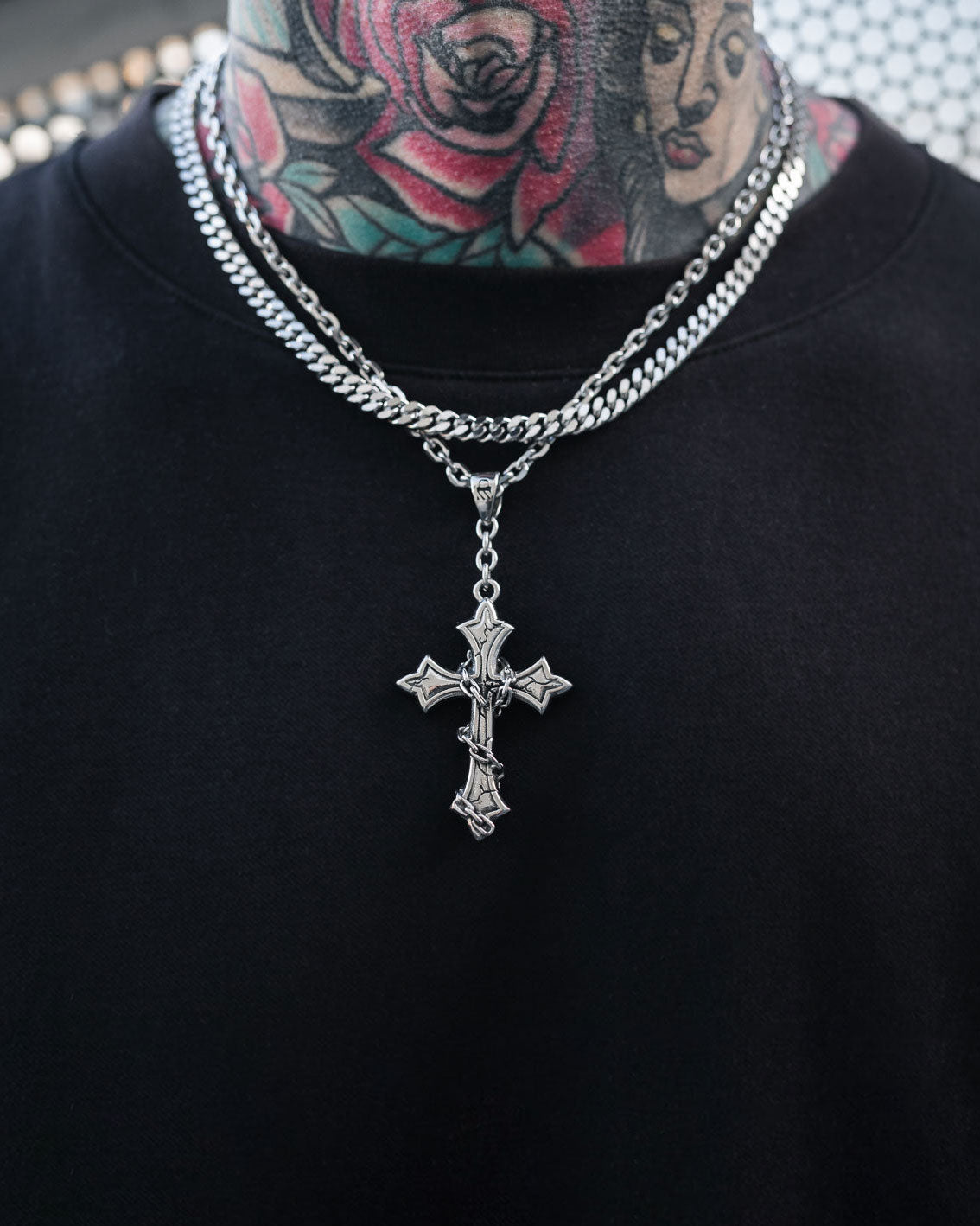 A person wearing a black shirt and Statement Collective's "The Ruins" 6mm Set, featuring layered stainless steel jewelry with a gothic cross pendant on a cuban link chain, showing off colorful floral and face tattoos on their neck.