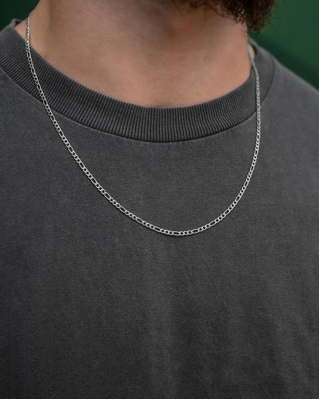 A close-up of a person wearing the Statement Collective 2.9mm Figaro Chain over a dark gray crew-neck t-shirt. The blurred green background highlights the necklace's non-tarnish shine.