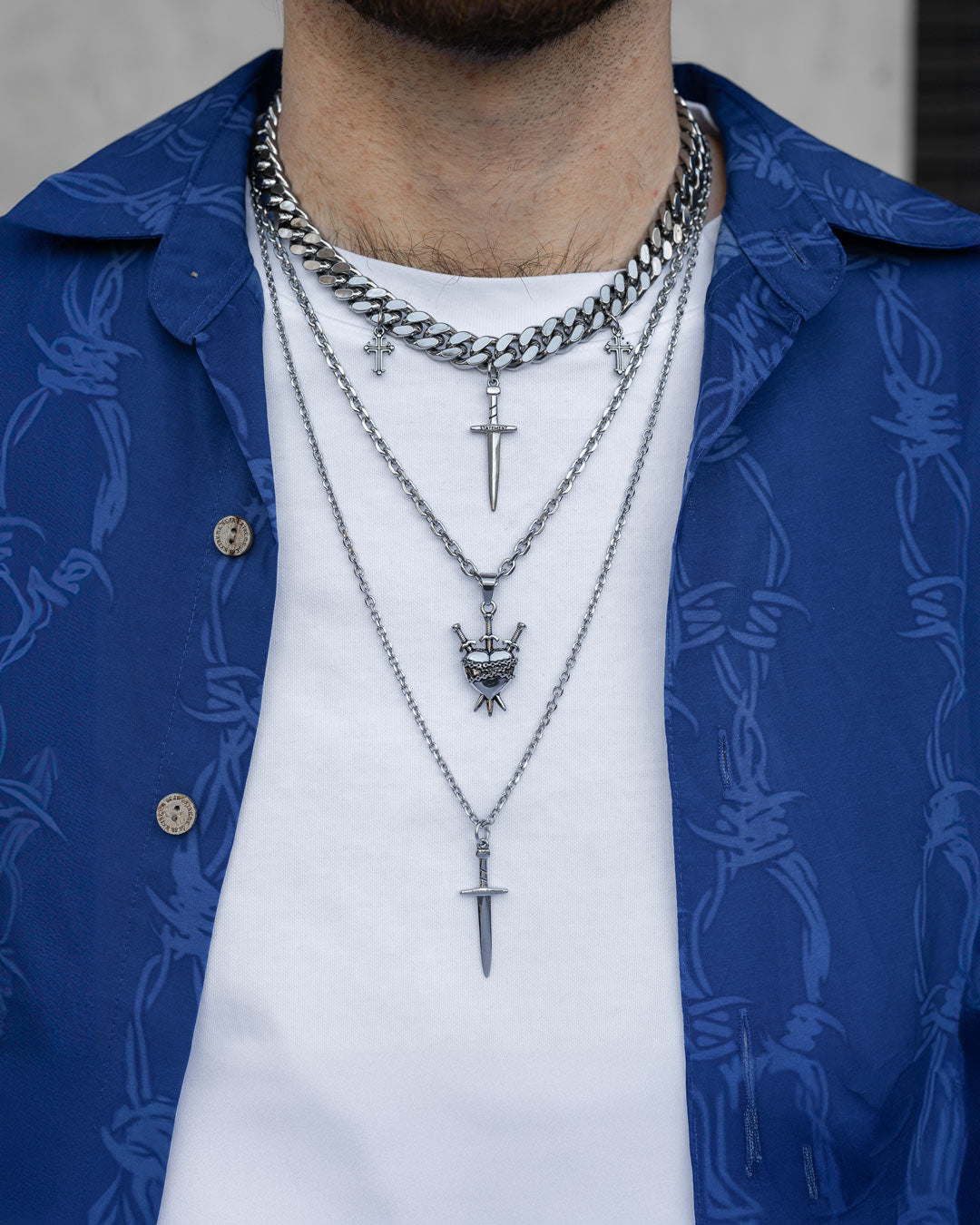 Someone wears the Angel & Dagger Stack, layering dagger pendants and a crown charm over a white t-shirt and an open blue shirt with a barbed wire pattern.