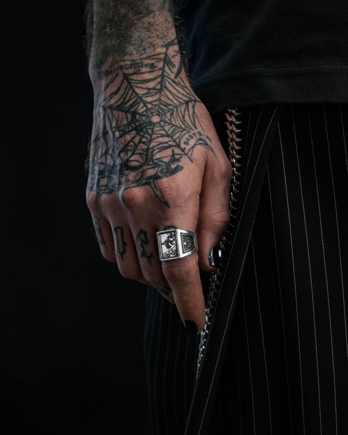 A tattooed hand adorned with the “Try Hard Fool” Ring by Statement Collective is displayed against dark, white-pinstriped clothing. The person’s fingernails are painted black and a spiderweb tattoo decorates the hand.