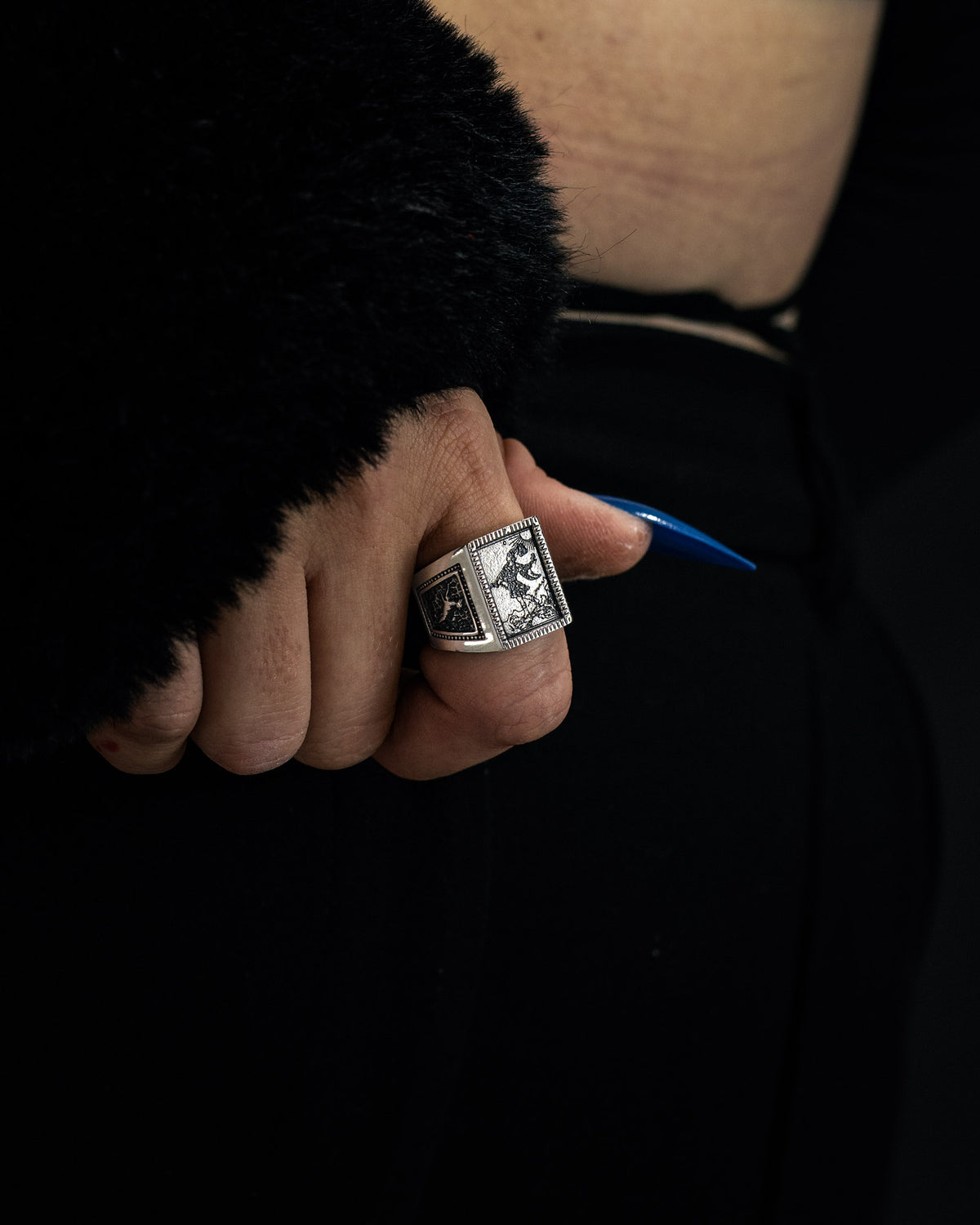 A hand with long blue nails rests on a hip, wearing the Statement Collective “Try Hard Fool” Ring—a large silver piece featuring an engraved design. The person is dressed in black, with a textured sleeve and a hint of skin visible at the waist.