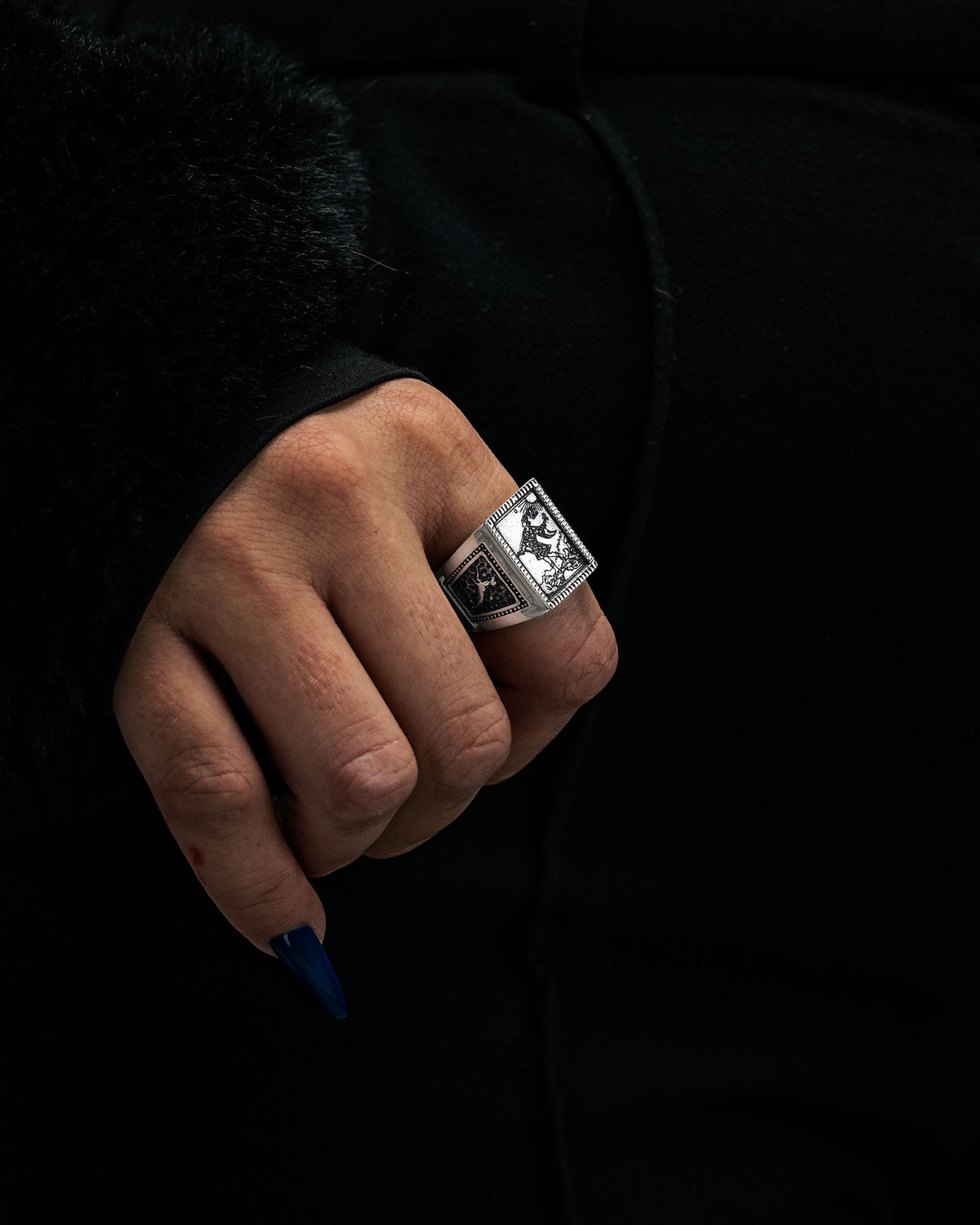 A hand with long blue nails wears Statement Collective’s “Try Hard Fool” ring, a large and ornate silver piece featuring intricate engraved designs. The person is dressed in black, with a visible furry sleeve edge.