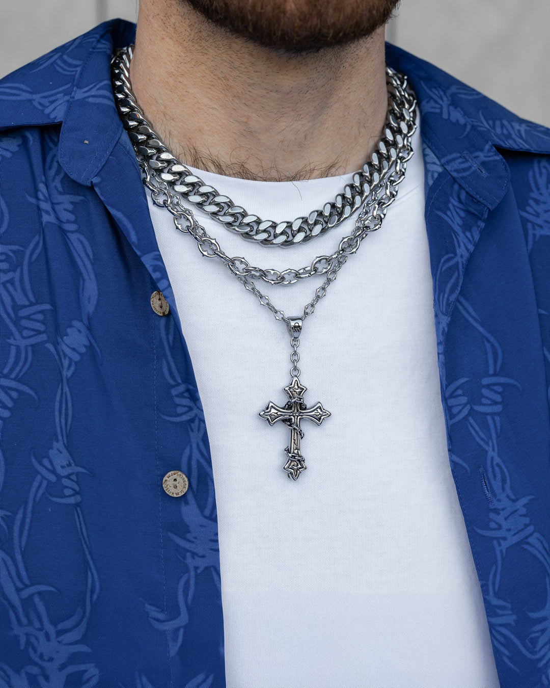 A person wearing the Untouchable Stack—layered silver chains featuring a Chunky Cuban chain and a cross pendant—with a white shirt and blue patterned overshirt. Only their lower face and neck are visible.