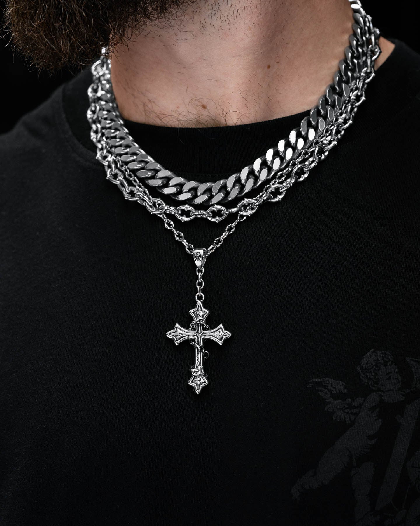 A close-up of a person wearing the Untouchable Stack—a chunky Cuban chain, silver cross pendant, and layered necklaces—over a black shirt with a subtle angel graphic on the shoulder. The background is black.