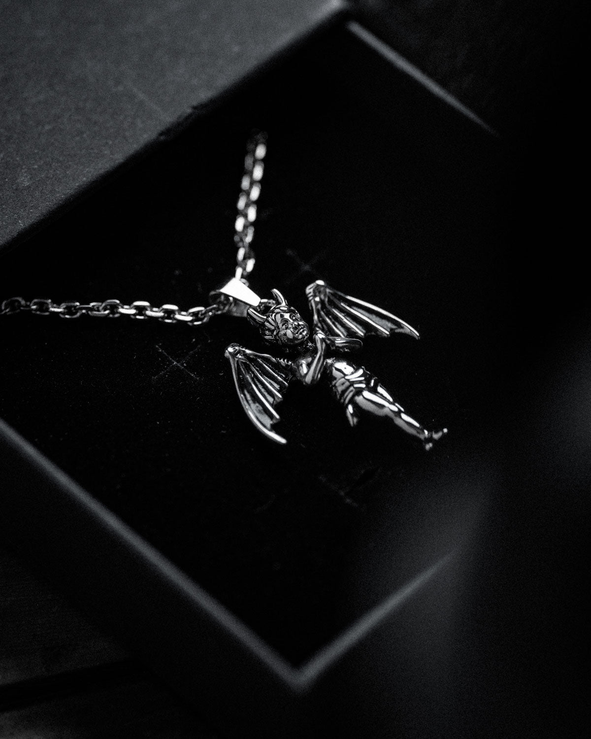 A black and white close-up of "The Wicked One" Pendant, a gothic necklace with a winged, horned cherub pendant, shown inside a black jewelry box to highlight its intricate details.