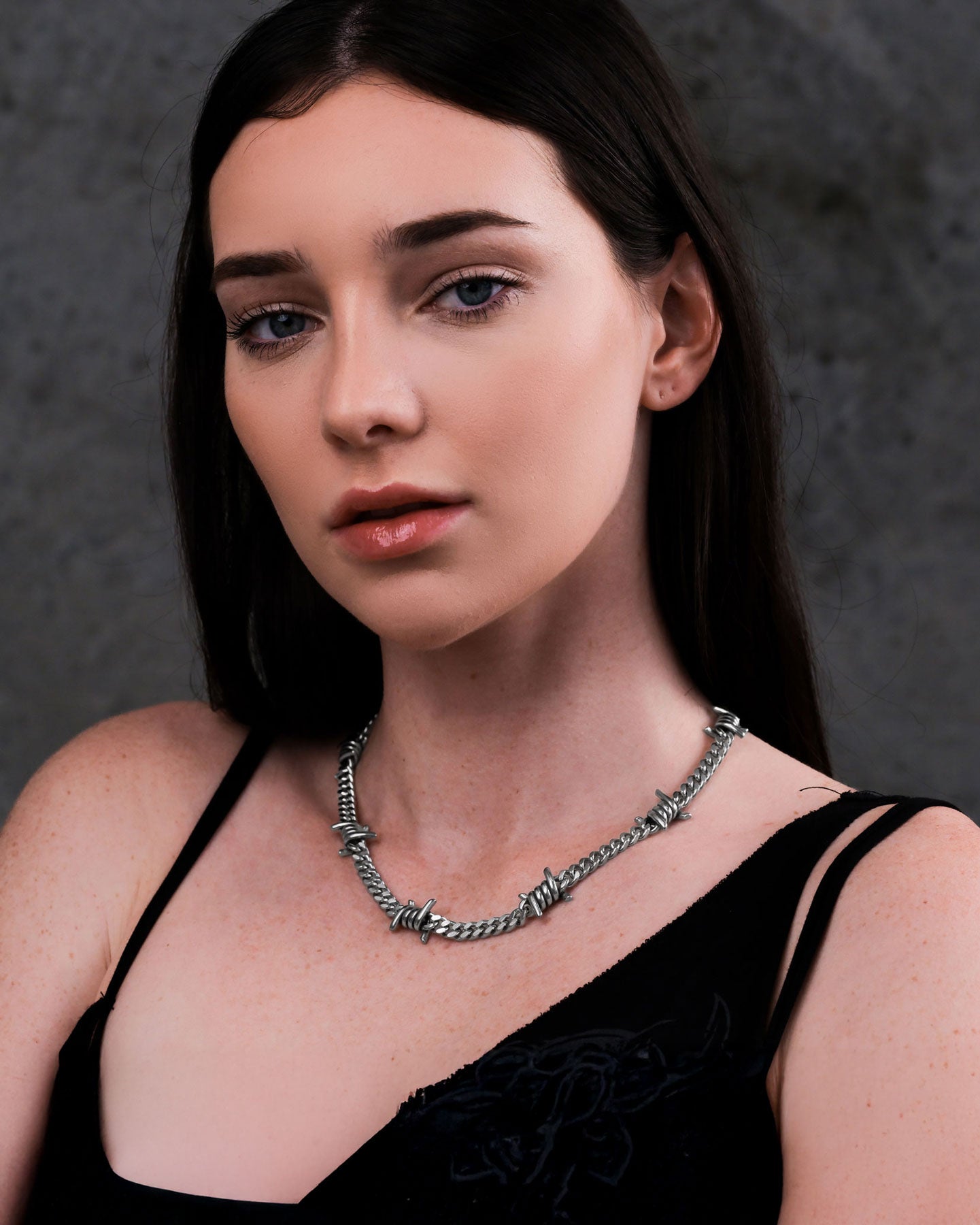 A woman with long dark hair and light skin wears a black top and the 6mm Barbed Wire Cuban Chain, posing against a dark, textured background that emphasizes her bold necklace.