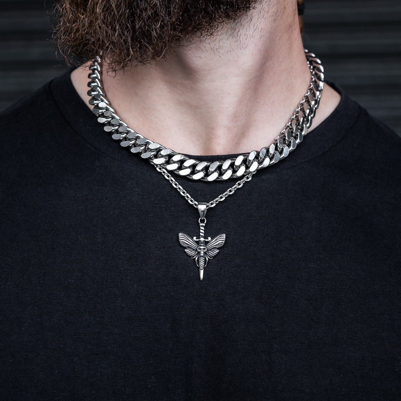 man with black shirt wearing death moth pendant necklace against dark wall